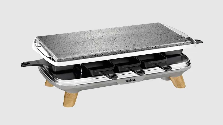 Een raclette-apparaat met een grijze steenplaat bovenop, zwarte pannetjes eronder en houten poten.