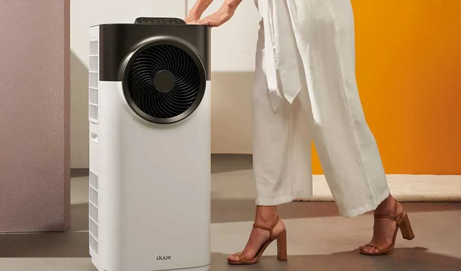 Een vrouw in witte kleding duwt een witte Duux airconditioner met groot zwart ventilatierooster; merk Duux duidelijk zichtbaar.
