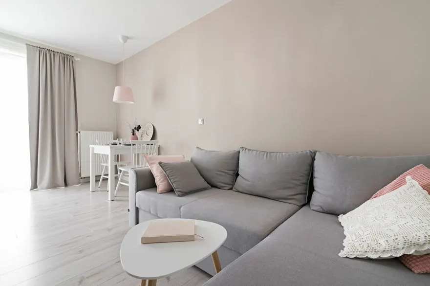 Een woonkamer met een grijze hoekbank met kussens, een witte tafel en stoelen, en grijze gordijnen.