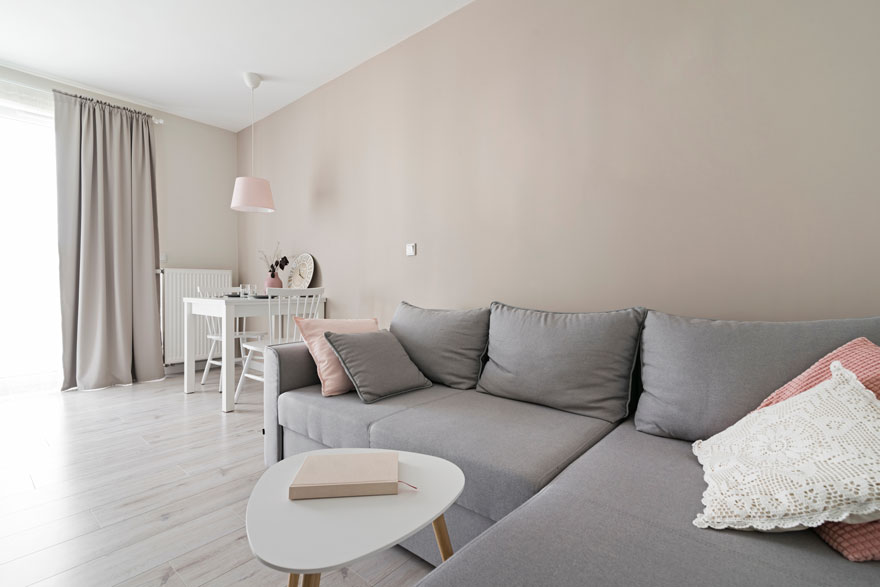 Salon beige avec canapé gris, coussins, table basse blanche, table et chaises blanches sous une lampe rose.