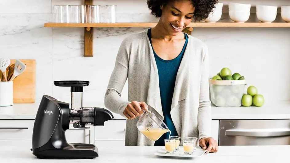 Une centrifugeuse noire à côté d'un pichet versant du jus dans des verres. Des pommes vertes sont visibles.
