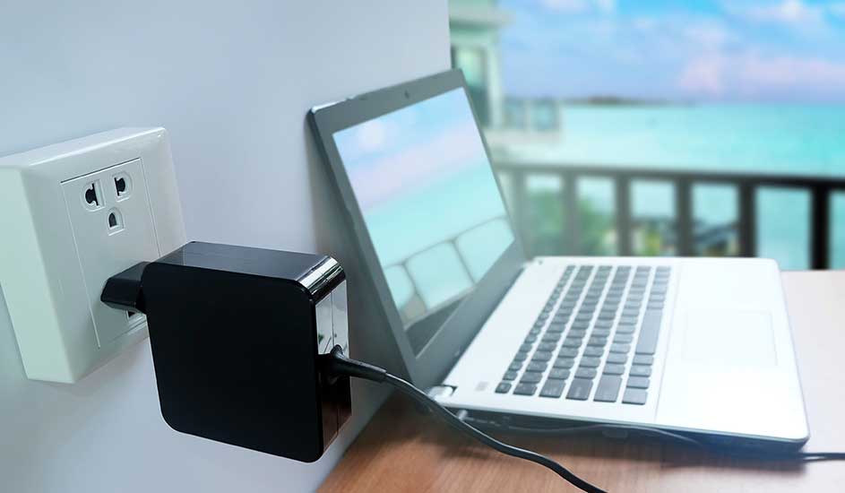 Een witte laptop staat op een tafel, aangesloten op een zwart adapterblok in een wit stopcontact tegen een witte muur. Op de achtergrond is een zeezicht.