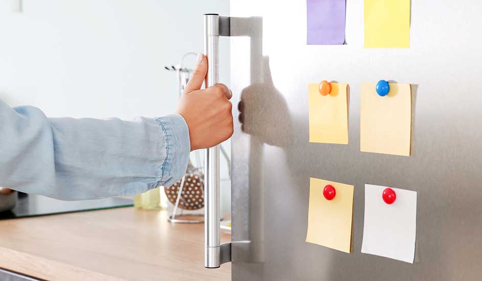 Een hand opent een zilverkleurige koelkast met gekleurde notities met punaises erop.