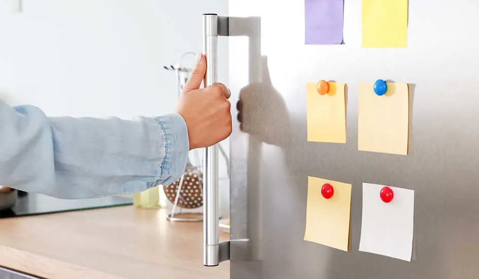 Een hand opent een zilverkleurige koelkast met gekleurde notities met punaises erop.