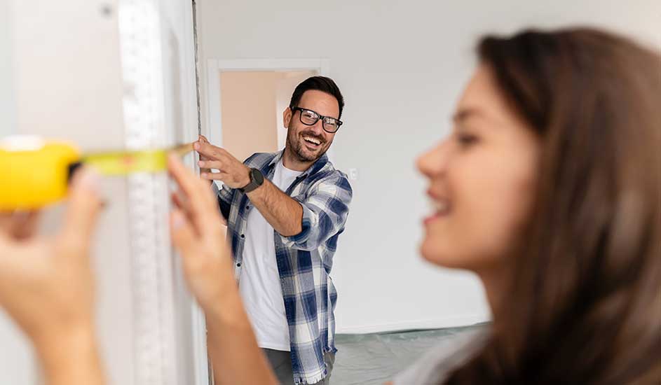 Un mètre ruban jaune mesure un mur blanc. Un homme en chemise à carreaux observe la mesure. L'arrière-plan est une pièce claire.