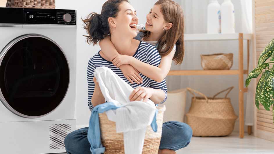 Une mère et sa fille souriant ensemble avec un panier à linge rempli de vêtements blancs et bleus. Un sèche-linge et une étagère en bois est visible à l'arrière.