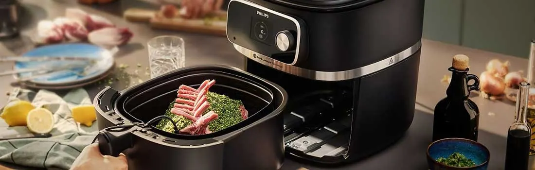 Black air fryer next to its basket containing lamb chops and herbs. Lemons, oil bottles, and onions in the background.
