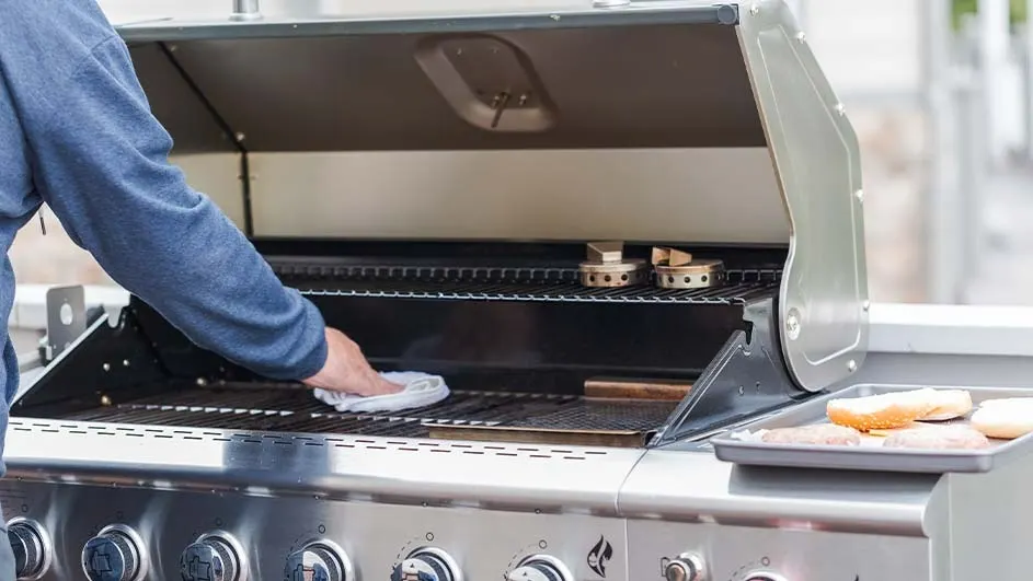 A stainless steel grill is being cleaned with a white cloth. The grill is open, revealing grates and accessories. A tray with food sits nearby.