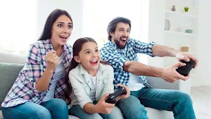 Een gezin zit op een grijze bank en speelt videospelletjes met zwarte controllers in een lichte kamer.