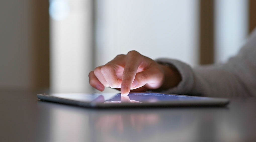 Een hand tikt op een helder tablet op een gladde tafel. De achtergrond is licht en onscherp.