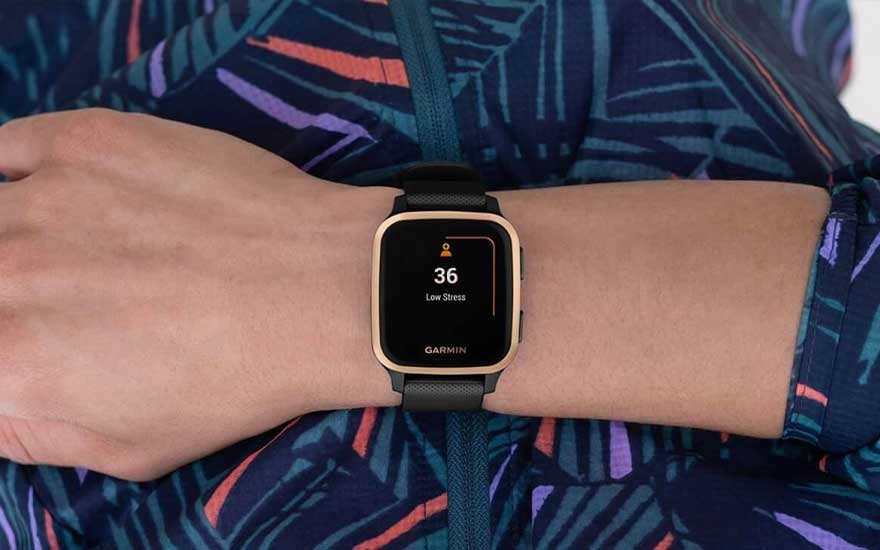 Une montre intelligente Garmin affiche « 36 » et « Stress faible » sur un poignet devant un tissu à motifs bleus, verts et violets.