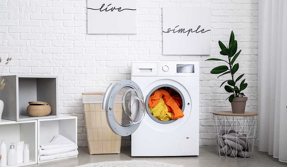 Witte wasmachine met open deur, gevuld met kleurrijke kleding, in een lichte kamer met witte bakstenen muur en decoratieve objecten.