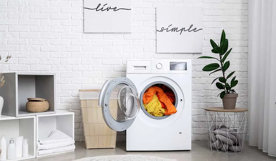 Witte wasmachine met open deur, gevuld met kleurrijke kleding, in een lichte kamer met witte bakstenen muur en decoratieve objecten.