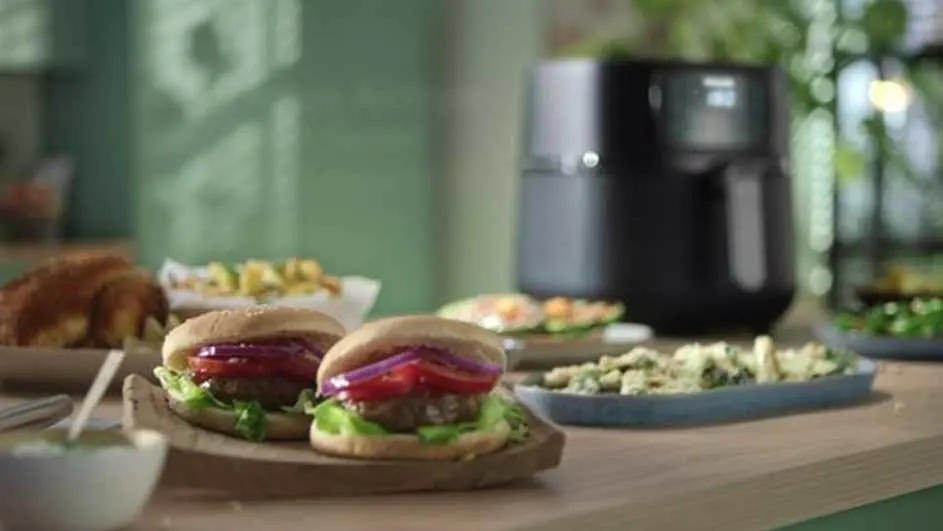 Deux hamburgers garnis de laitue, tomate et oignon rouge sur une table en bois avec divers plats et une friteuse à air noir en arrière-plan.