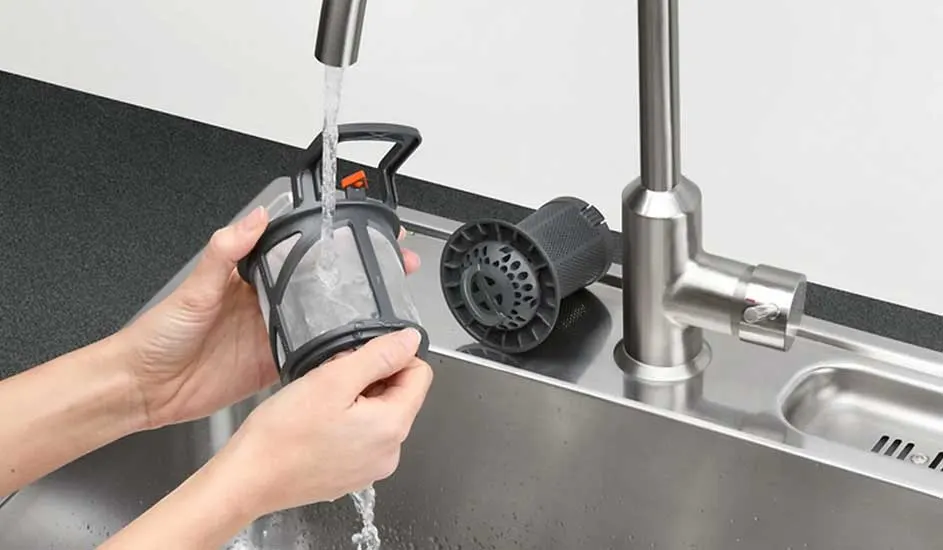 A person rinses a dishwasher filter under running water at a metal sink. No brand is visible on the filter.
