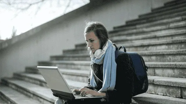 Une personne assise sur des marches en béton utilise un ordinateur portable, portant un foulard bleu et un sac à dos.
