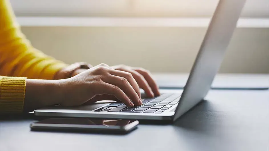 Een persoon typt op een slanke laptop aan een bureau, met een smartphone ernaast en zacht daglicht op de achtergrond.