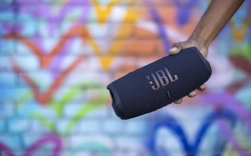 Haut-parleur JBL noir tenu devant un mur coloré avec des motifs en V en orange, bleu, jaune et vert.