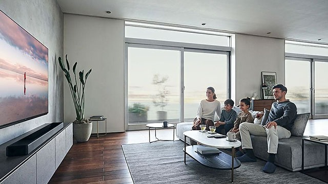 Salon lumineux avec TV grand écran, canapé gris, table basse et grandes fenêtres coulissantes.