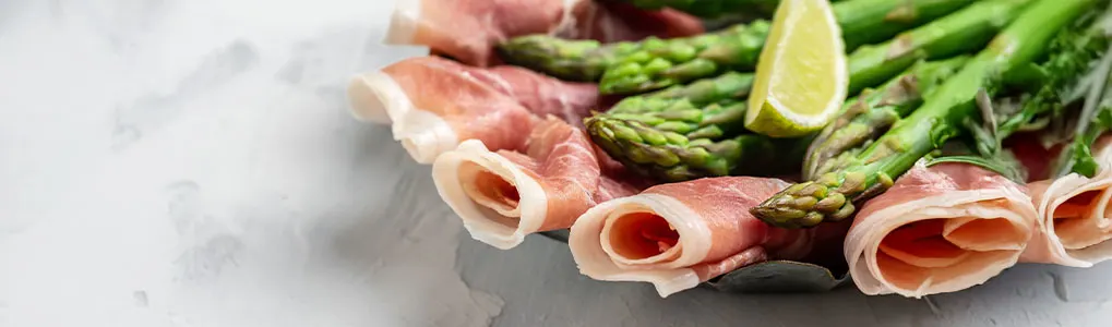 Assiette avec asperges vertes, jambon roulé et citron vert sur un fond gris clair.