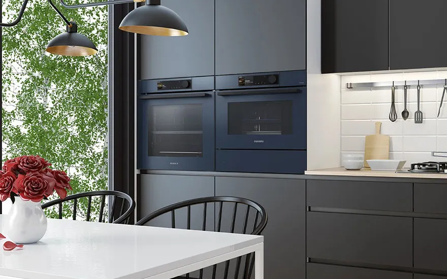 Moderne keuken met blauwe ovens, grijze kasten, wit aanrecht en tafel. Rode rozen in een vaas. Keukengerei hangt aan de muur.