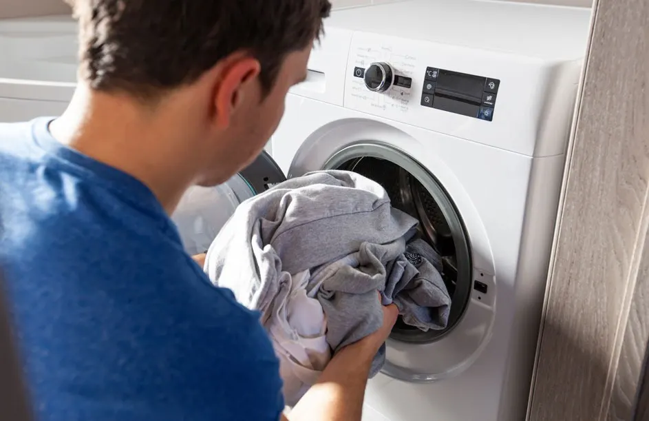 Een witte wasmachine met grijze kleding erin. De machine heeft een draaiknop en een digitaal scherm.