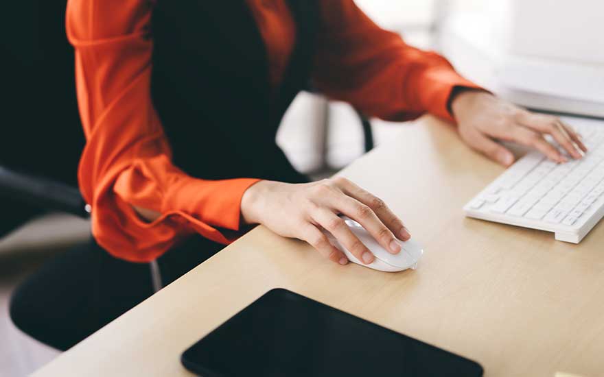 Une femme en blouse orange travaille à son bureau, utilisant une souris blanche et un clavier, une tablette à côté.