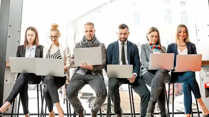 Five people sit in a row with laptops. They wear business casual clothing. A building is visible through the window.