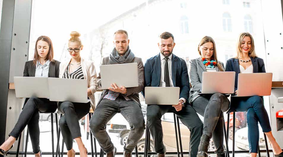 Vijf mensen zitten op krukken met laptops. Achter hen is een groot raam met een gebouw zichtbaar.