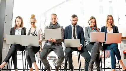 Vijf mensen zitten op krukken met laptops. Achter hen is een groot raam met een gebouw zichtbaar.