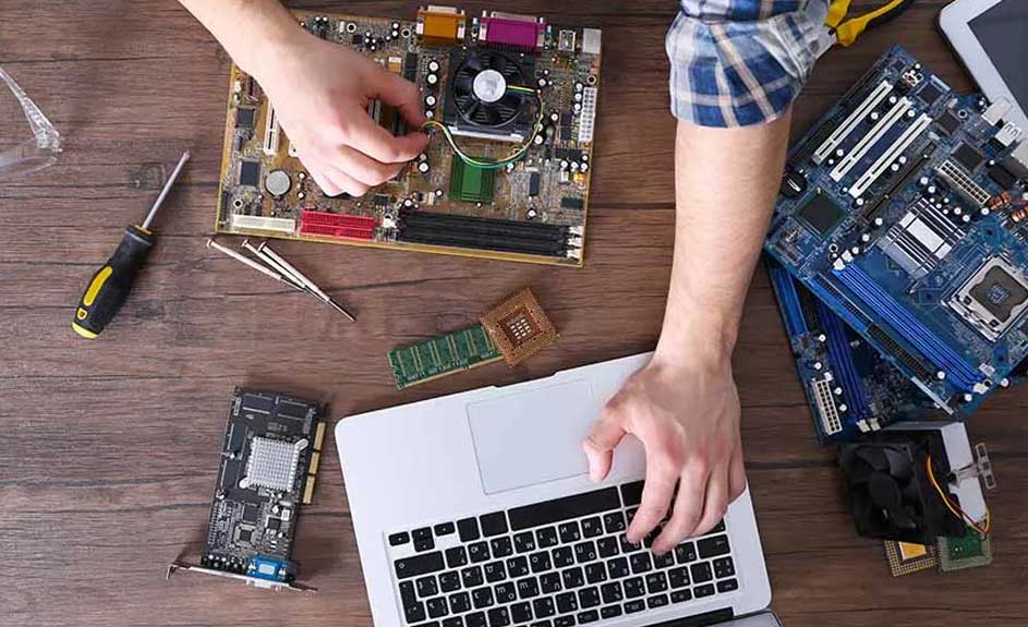 Een houten tafel met een laptop, printplaten, gereedschap en computeronderdelen erop. Iemand typt op de laptop.
