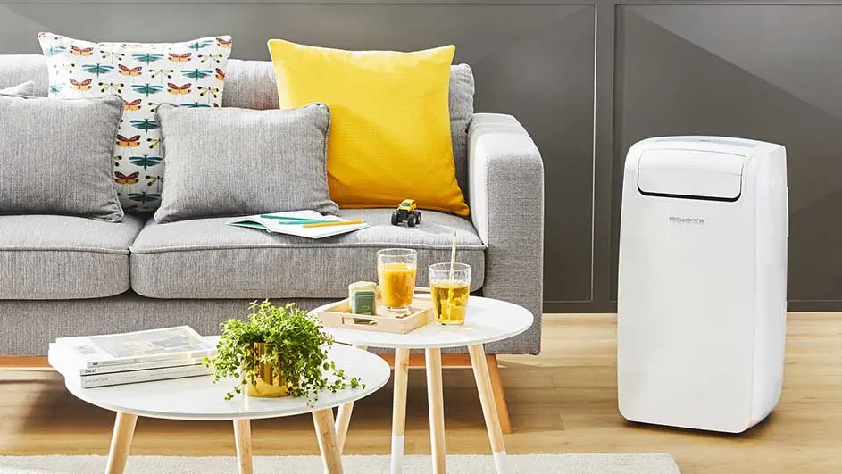 Woonkamer met grijze sofa, gele kussen, tafels met drankjes, plant en witte airco tegen een grijze wand.
