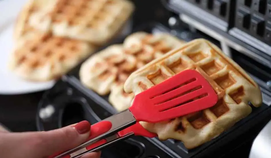 Een wafel in een wafelijzer wordt met een rode tang verwijderd. Achtergrond: bord met wafels.