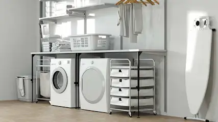 Een wasruimte met een wasmachine, droger, strijkplank en opbergrekken in wit en grijs.