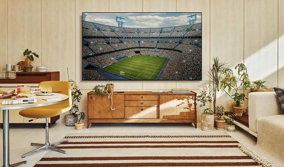 Une grande télévision montre un stade de football plein dans un salon lumineux avec meuble en bois, plantes, table et douce lumière.