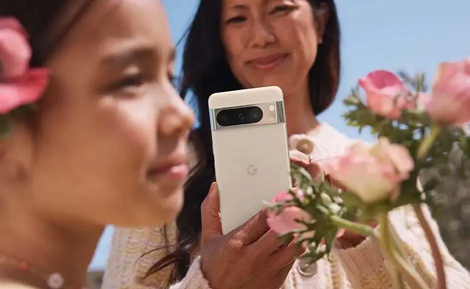 Une personne tient un téléphone blanc avec un bloc de caméra noir, entouré de fleurs roses. Ciel bleu en arrière-plan.
