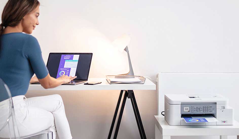 Bureau blanc avec ordinateur portable, lampe et imprimante. L'écran affiche des graphiques. Imprimante blanche avec papier bleu.