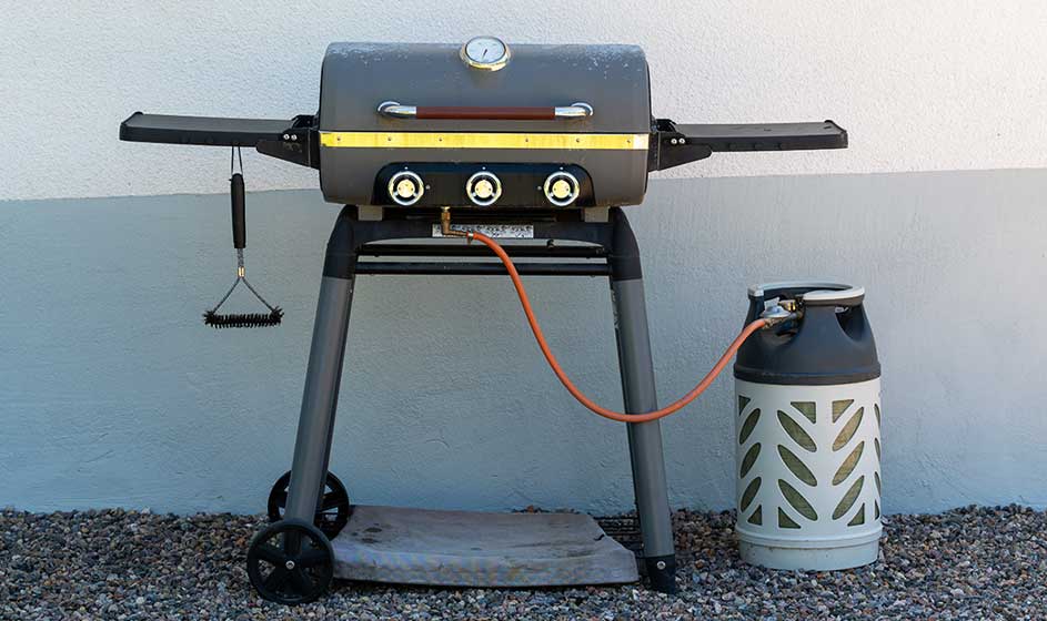 Gasbarbecue met zijplanken staat klaar naast een gasfles; een tang hangt links en de slang verbindt beide toestellen.