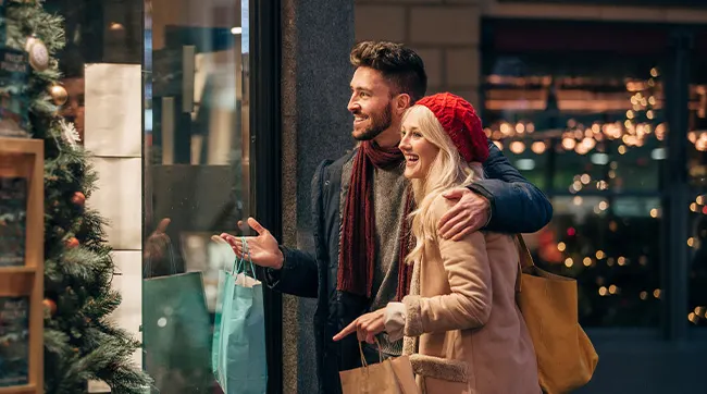 Couple avec écharpes et sacs de shopping souriant devant une vitrine décorée.