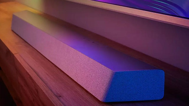 Une barre de son rectangulaire est posée sur une surface en bois, sous un écran de télévision. L'éclairage est violet et bleu.