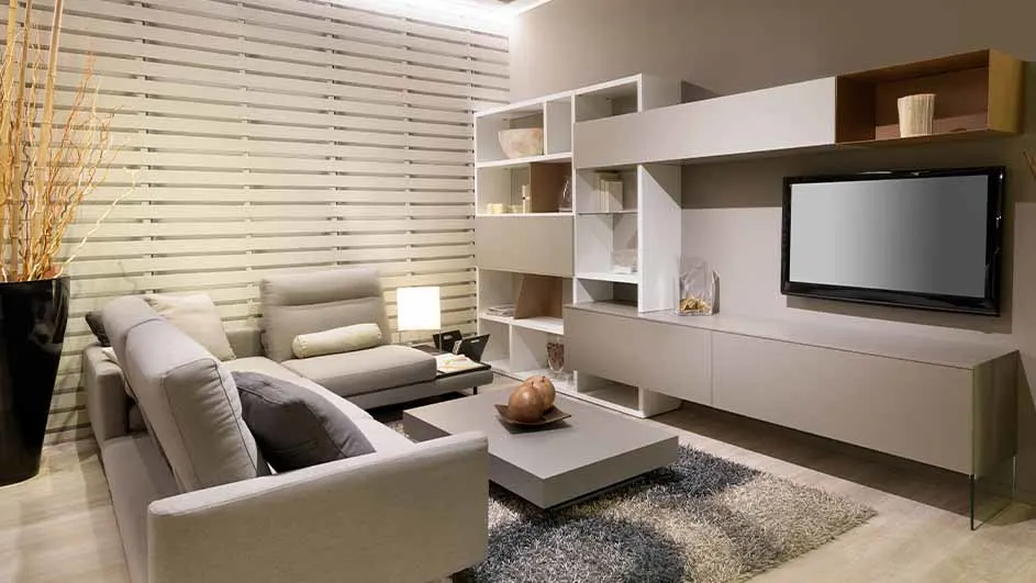 Salon beige avec canapé, table basse, TV et étagères. Un vase noir est visible à gauche.