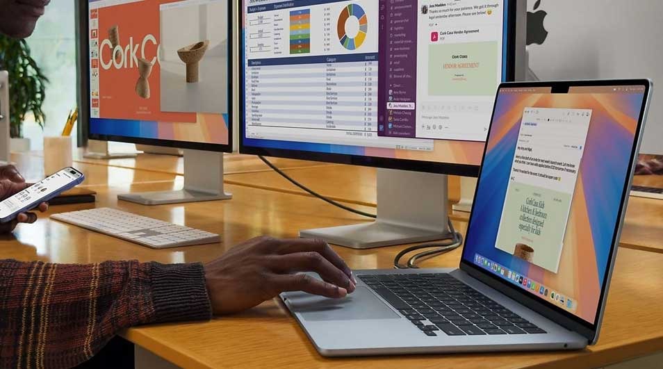 A person works on an Apple MacBook linked to two large monitors, while holding an iPhone, on a wooden desk with keyboards and tidy cables.