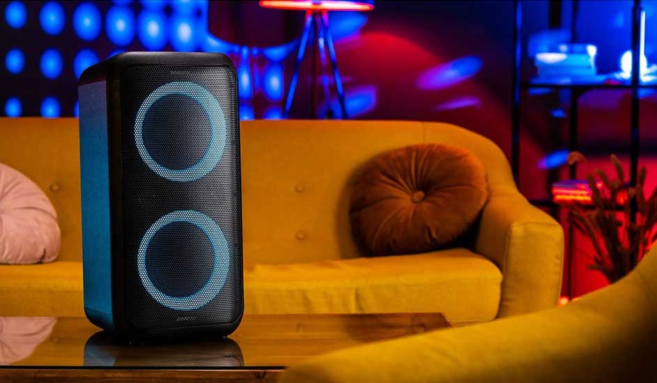 Dit is een grote zwarte Sony partyspeaker met blauwe lichtcirkels, geplaatst op een tafel voor een gele bank in een kleurrijke kamer.