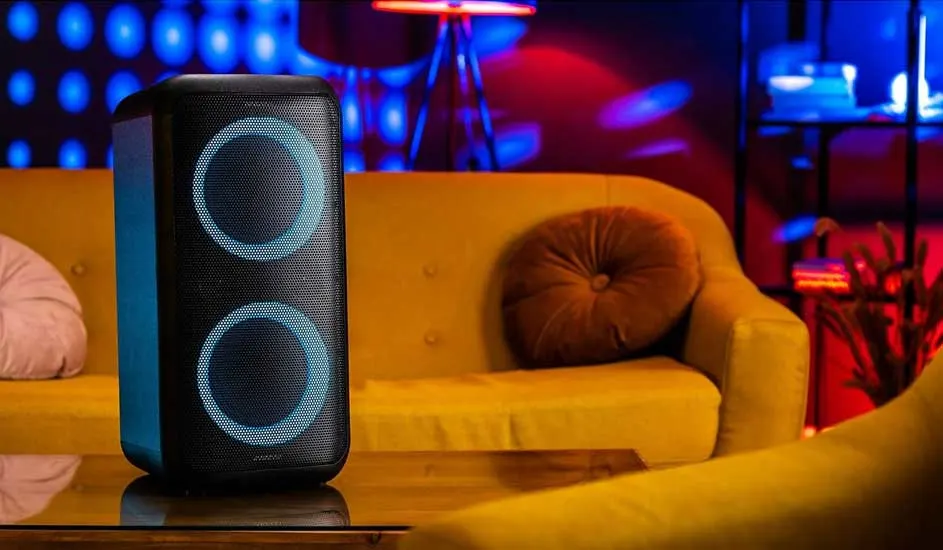 Voici une grande enceinte Sony noire avec des cercles lumineux bleus, posée sur une table devant un canapé jaune dans une pièce colorée.