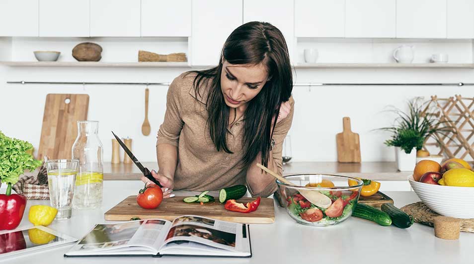 Een keuken met een houten snijplank waarop groenten worden gesneden, naast een kookboek en ingrediënten voor een salade.