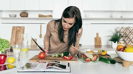 Een keuken met een houten snijplank waarop groenten worden gesneden, naast een kookboek en ingrediënten voor een salade.
