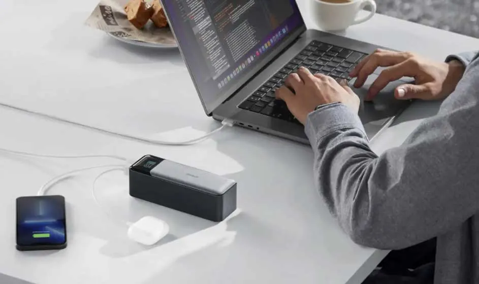 Une personne travaille sur un ordinateur portable. À côté se trouvent un smartphone, un chargeur, un câble, des écouteurs et une tasse de café.