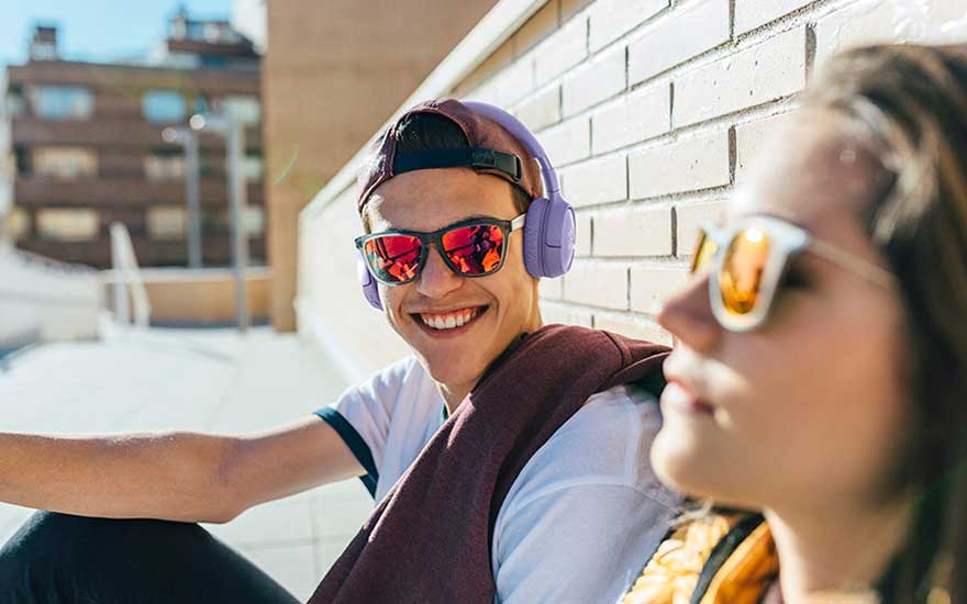 Un jeune homme porte un casque violet et des lunettes de soleil rouges, assis devant un mur de briques.