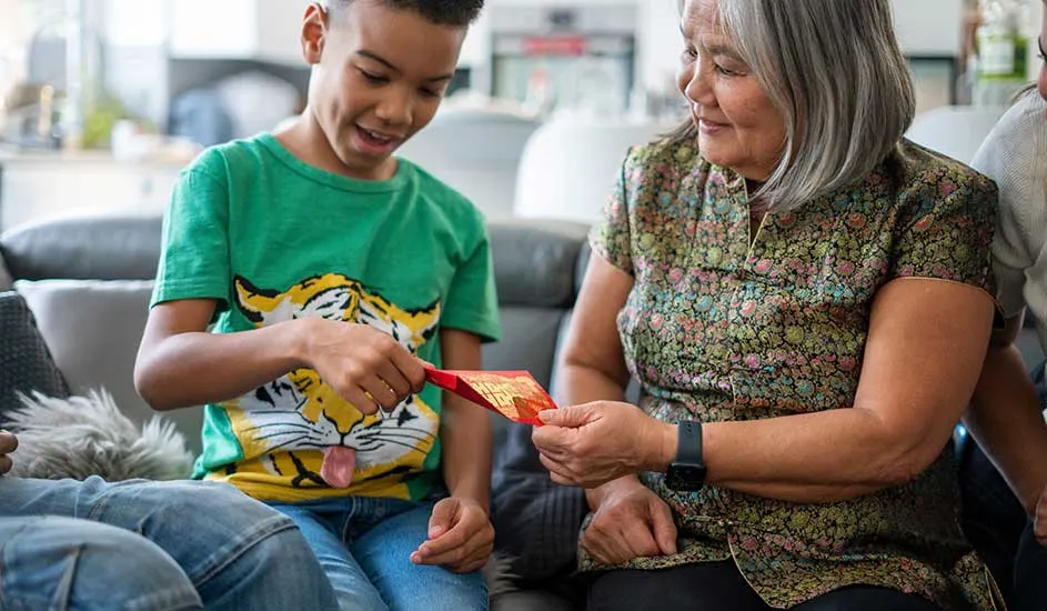 Un enfant en t-shirt tigre reçoit une enveloppe rouge d'une personne en chemisier à fleurs. Le tout se passe dans un salon.