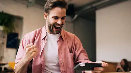 Un homme en chemise rose tient un téléphone. Il a le poing levé et sourit, sur fond d'un intérieur de café lumineux.